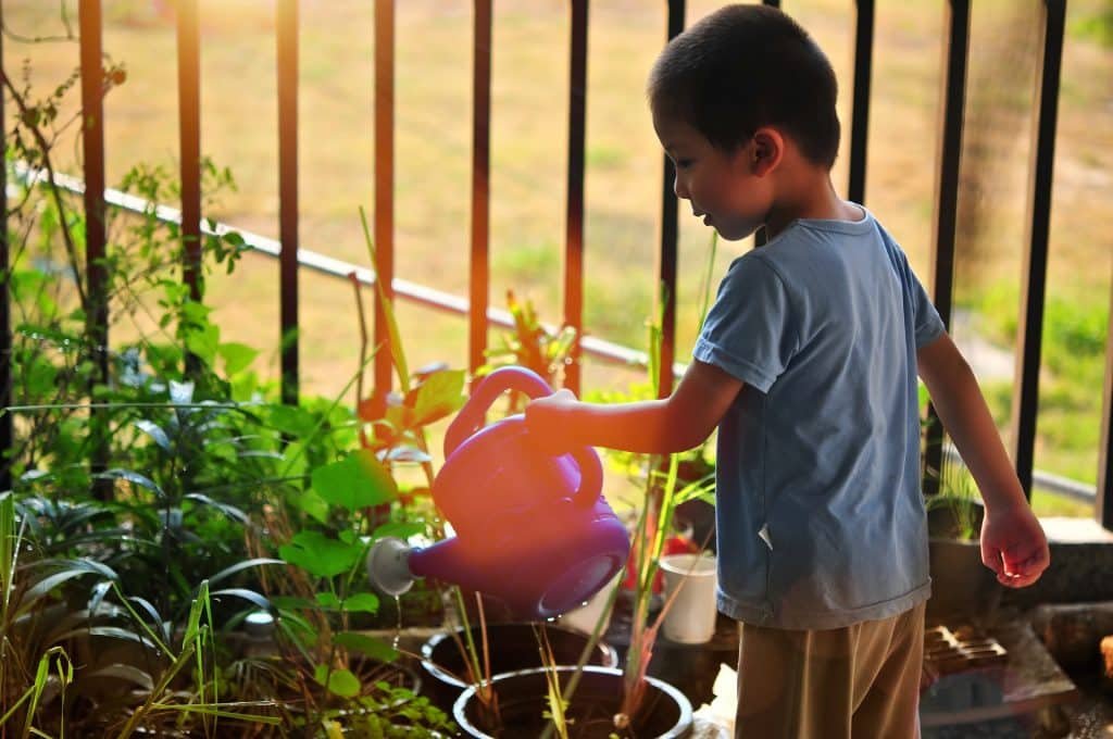 Een jong kind in een blauw shirt gebruikt tuingereedschap om planten water te geven op een balkon met een blauwe gieter. De zon werpt een warme gloed en potplanten zijn gerangschikt voor een reling met gras zichtbaar op de achtergrond, wat een ideaal tafereel creëert voor kinderen.