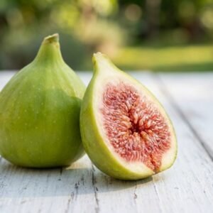An unrooted fig cutting 'Col de Dame Blanc' is displayed on a white wooden surface, with a whole green fig and a half-cut fig showing its pink interior, set against a blurred green garden background.